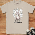 A beige graphic t-shirt laid flat on a wooden surface, featuring a white cat surrounded by small white ghosts and the text '9 LIVES 0 REGRETS'. Black sneakers are partially visible in the bottom right corner.