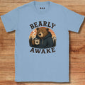 Light blue t-shirt with a bear graphic and 'BEARLY AWAKE' text on a wooden surface