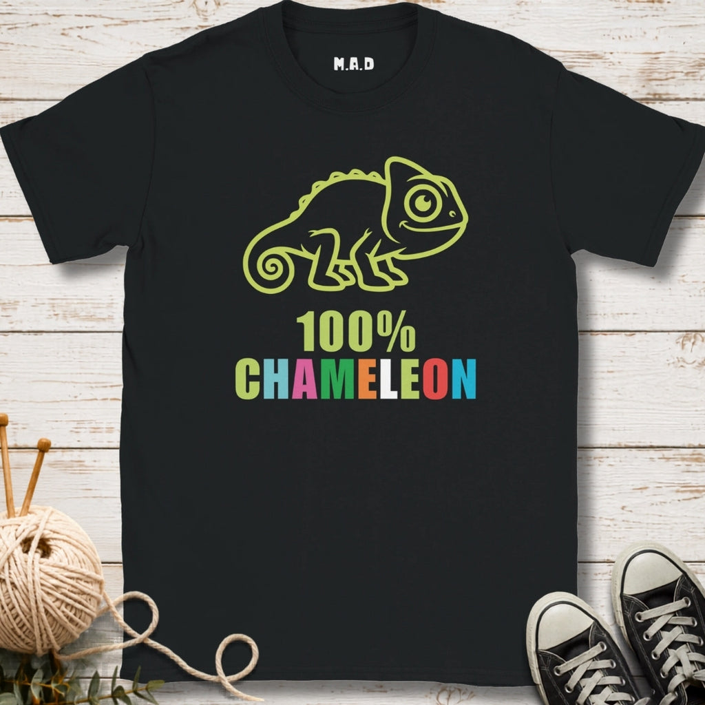 A black short-sleeve graphic T-shirt laid flat on a whitewashed wood surface, featuring a lime green chameleon outline above the words '100% CHAMELEON' in multicolored letters.