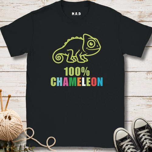 A black short-sleeve graphic T-shirt laid flat on a whitewashed wood surface, featuring a lime green chameleon outline above the words '100% CHAMELEON' in multicolored letters.