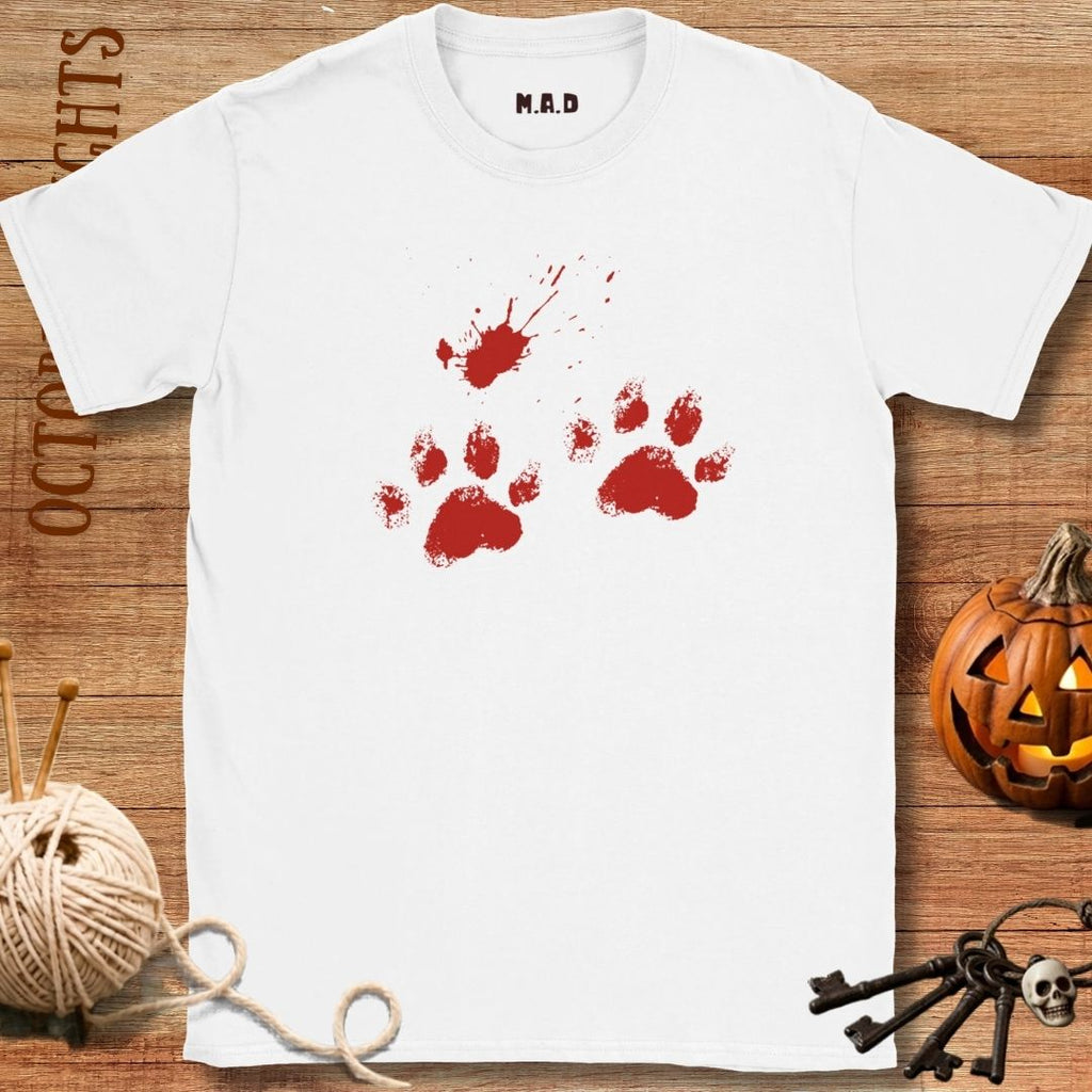 A white short-sleeve T-shirt laid flat on a wooden surface, featuring a red paw-print and blood-splatter graphic on the chest. The shirt is styled with Halloween props including a jack-o'-lantern, skeleton keys, a ball of yarn, and a spider decoration.