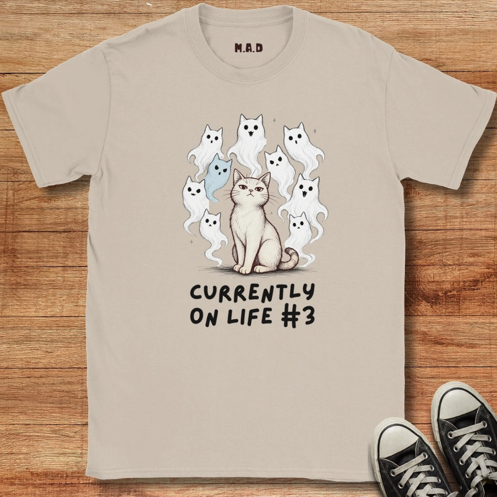 Beige t-shirt with a graphic of a cat and ghosts and text 'Currently on life #3' on a wooden surface.
