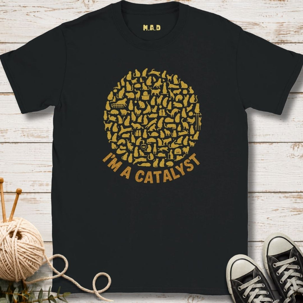 A black short-sleeve graphic T-shirt laid flat on a white wood plank background, with a large gold circular chest print made of small tool icons and the words 'I'M A CATALYST' below. A small gold 'M.