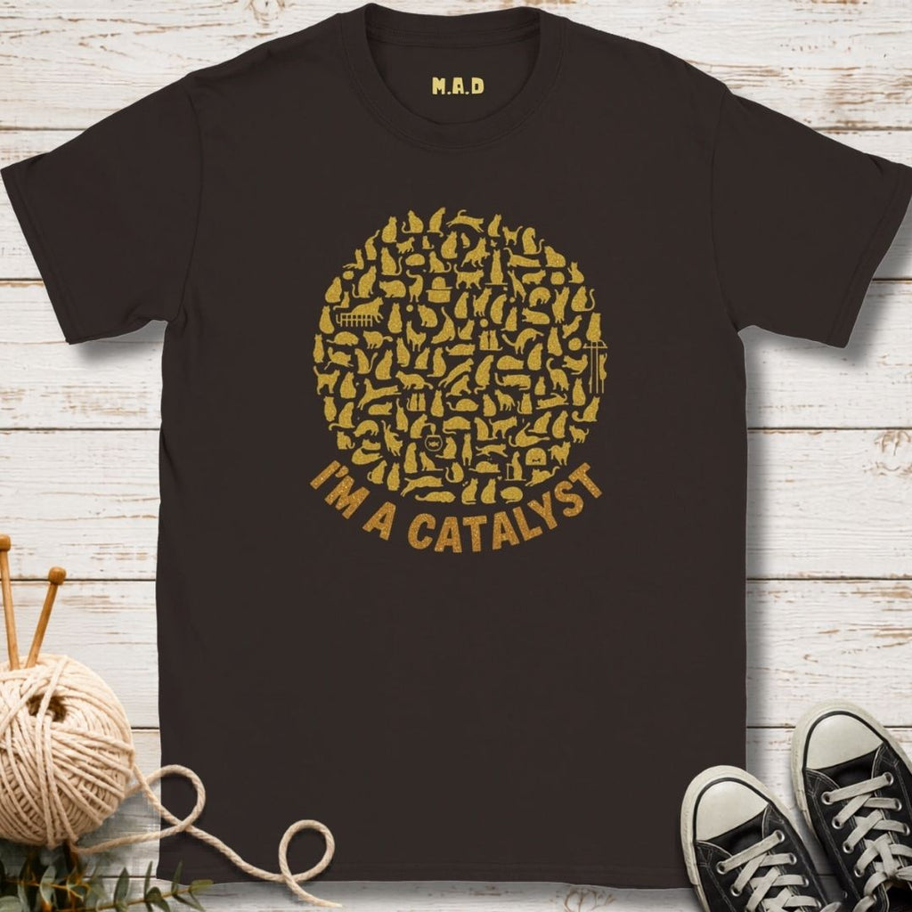 A dark brown crewneck t-shirt with a gold graphic on the front. The graphic features a circle filled with knitting needles and yarn icons, with the text 'I'M A CATALYST' printed below in gold.
