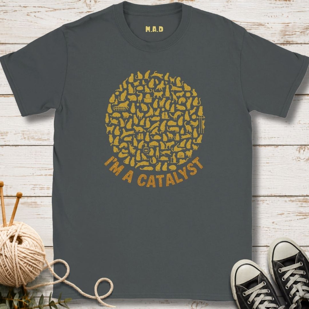 A dark gray t-shirt with a gold graphic of knitting needles forming a circle and the text 'I'M A CATALYST' underneath. The shirt is displayed flat on a white wood background with decorative props including a ball of yarn, knitting needles, and black sneakers.