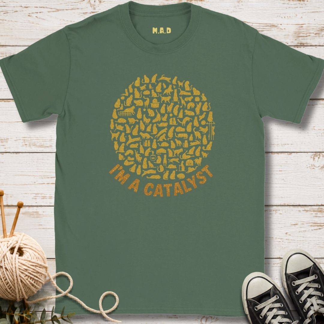 A sage green t-shirt with a yellow graphic of knitting needles forming a circle and the text 'I'M A CATALYST' underneath. The shirt is displayed flat on a white wood background with decorative props including a ball of yarn, knitting needles, and black sneakers.