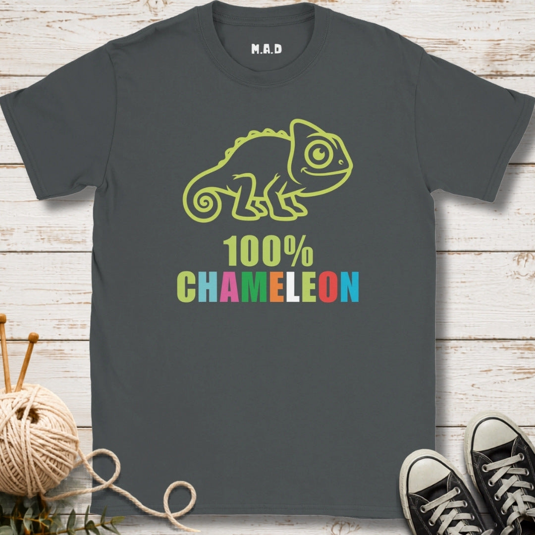 A dark gray t-shirt with a lime green chameleon graphic and the text '100% CHAMELEON' in multicolor letters, displayed on a white wood background with knitting props and sneakers.