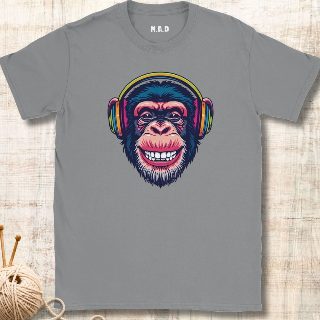 A gray short-sleeve crewneck t-shirt featuring a colorful graphic of a smiling chimpanzee wearing headphones, displayed flat on a light wood background with a ball of yarn and knitting needles in the corner.