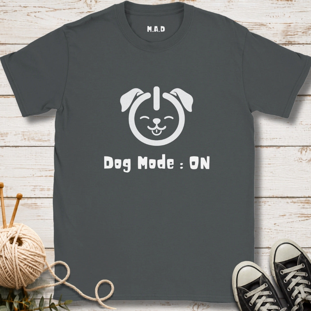 A dark gray t-shirt with a white graphic of a dog head and the text 'Dog Mode : ON' printed on the front. The shirt is displayed in a flat-lay arrangement on a white wood background, styled with a ball of yarn, knitting needles, and black sneakers.