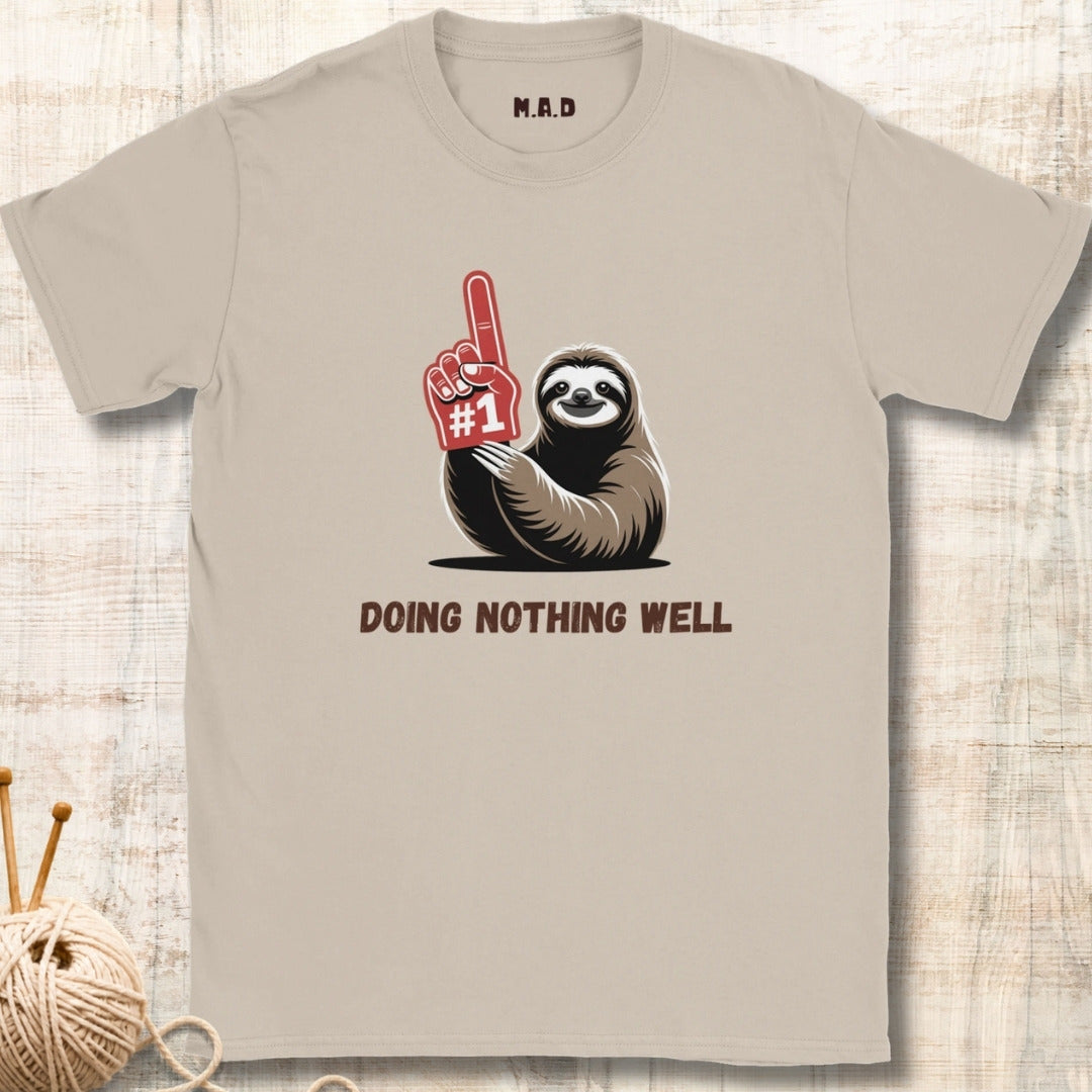 Beige t-shirt with a sloth holding a foam finger and text 'Doing Nothing Well' on a wooden surface.