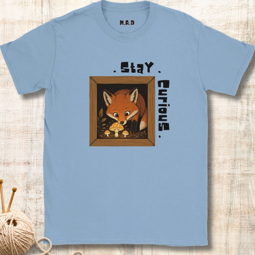 A light blue crewneck t-shirt with a graphic of a cute cartoon fox in a wooden frame looking at mushrooms, with the text 'Stay Curious' printed around it. The t-shirt is displayed flat on a light wood background with a ball of yarn and knitting needles in the corner.