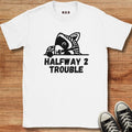 A white short-sleeve graphic T-shirt laid flat on a wooden surface, featuring a black cartoon raccoon peeking over the text 'HALFWAY 2 TROUBLE.' Black sneakers are partially visible at the bottom right as styling props.