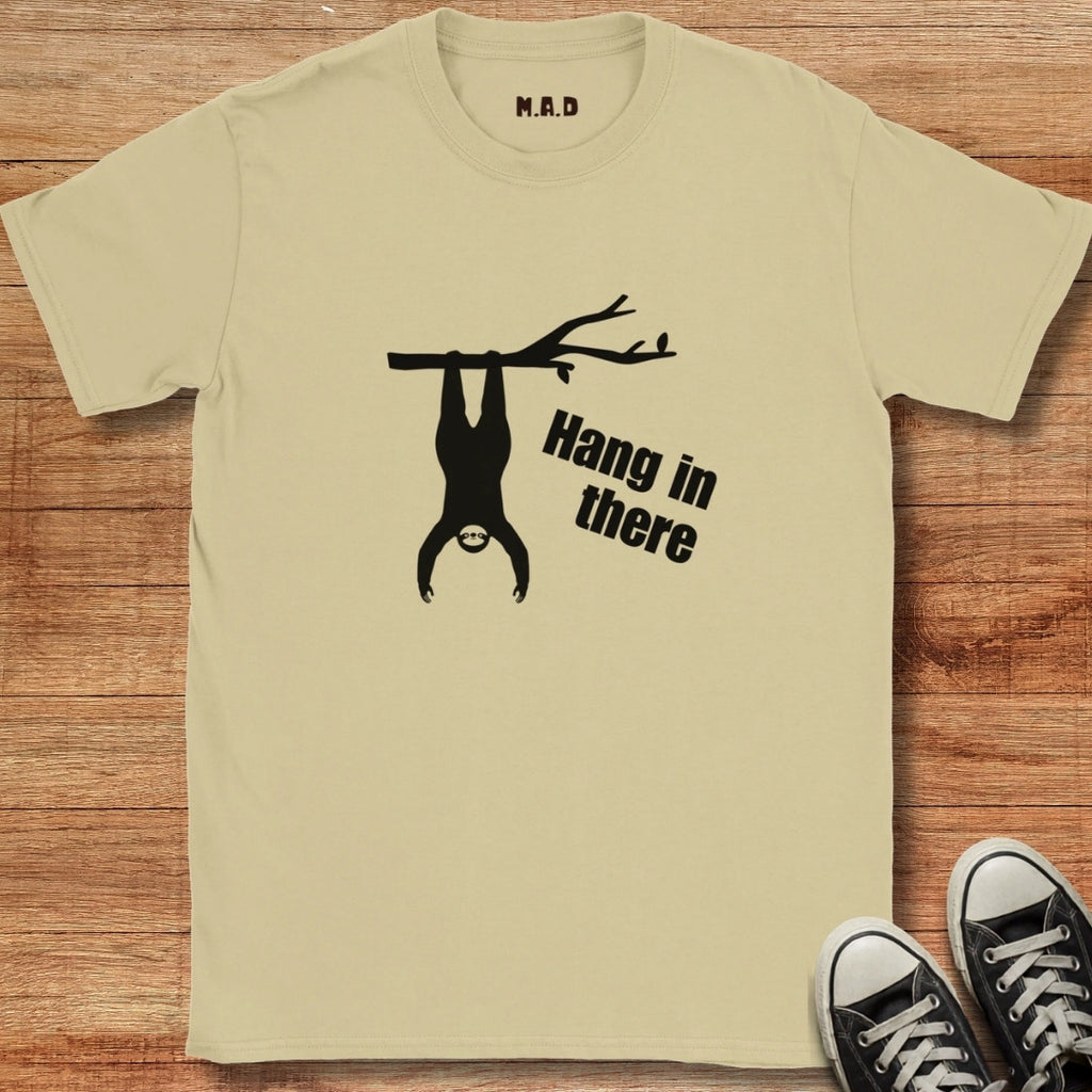 Beige t-shirt with a sloth graphic and 'Hang in there' text on a wooden surface, next to black sneakers.