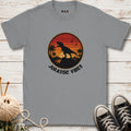 Gray t-shirt with 'Jurassic Vibes' design featuring a dinosaur silhouette, placed on a wooden surface with sneakers and a ball of yarn.