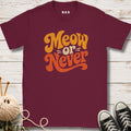 A maroon short-sleeve t-shirt with a retro-style graphic reading 'Meow or Never' in orange and yellow lettering, laid flat on a white wood plank background with knitting props and black sneakers nearby.