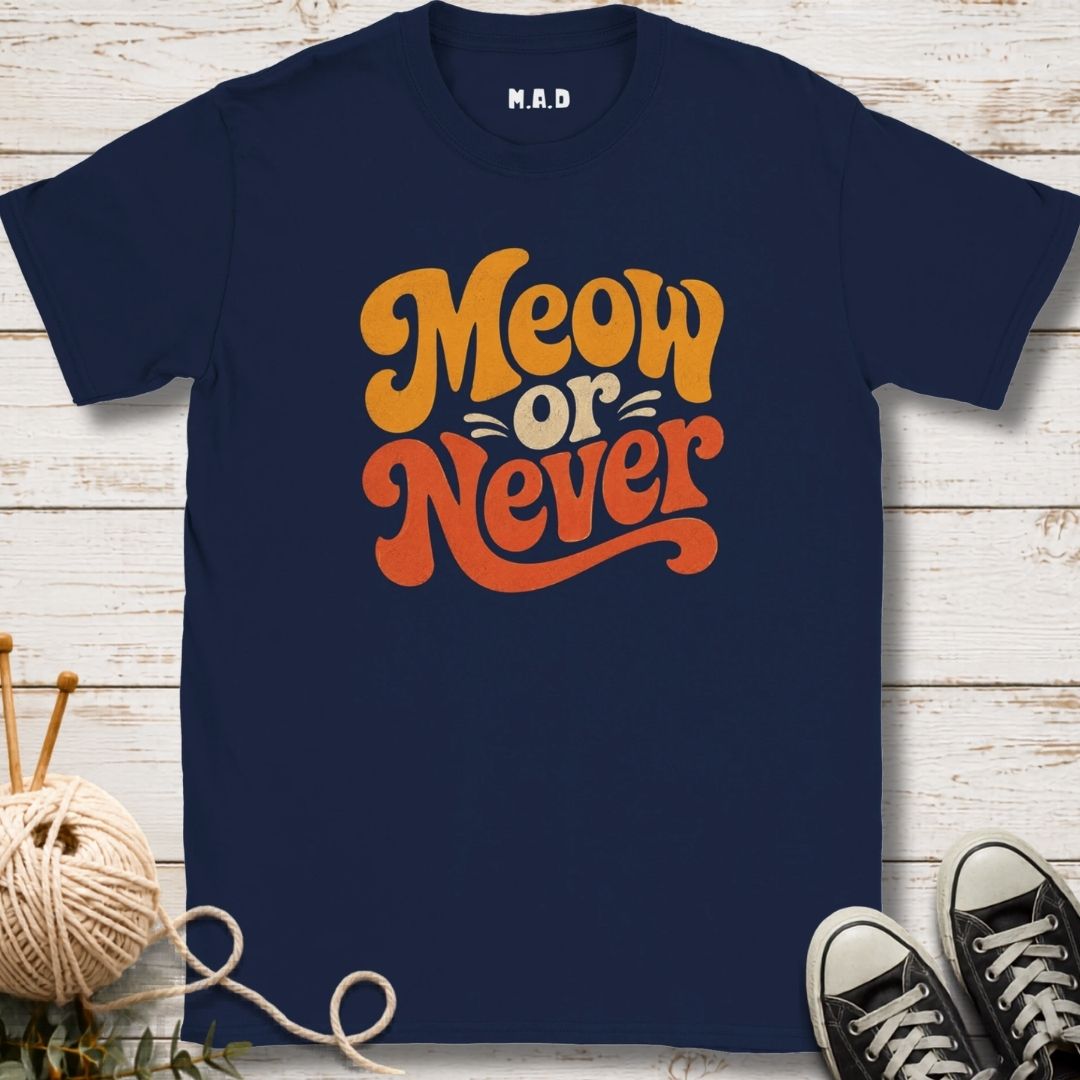 A navy blue short-sleeve t-shirt with a retro-style graphic reading 'Meow or Never' in orange and yellow lettering, displayed on a white wood plank background with knitting props and black sneakers.