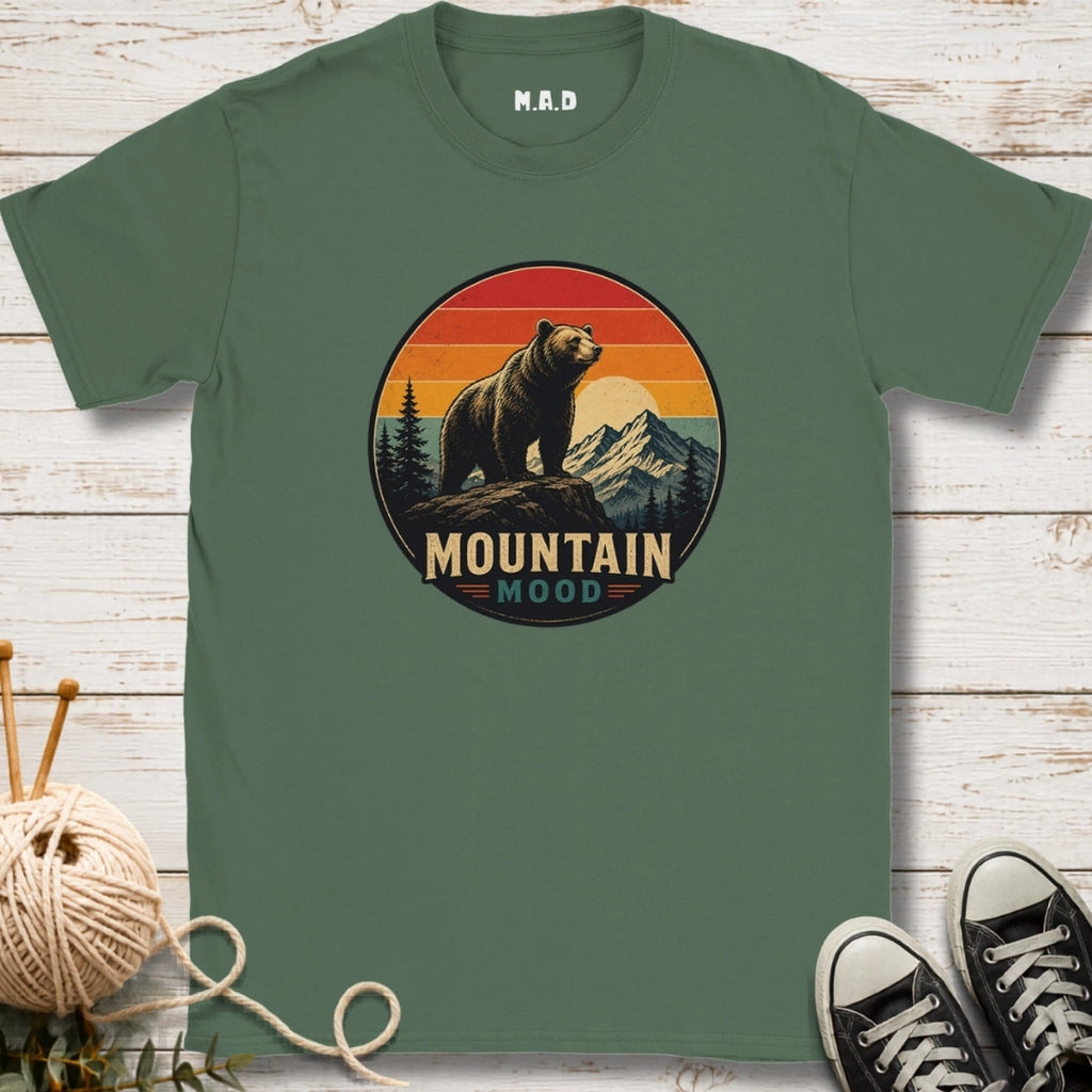 A sage green short-sleeve t-shirt with a circular retro graphic of a bear in front of mountains and a sunset, with the text 'MOUNTAIN MOOD'. The t-shirt is displayed flat on a white wood background with knitting needles and yarn on the left, and black sneakers on the right.