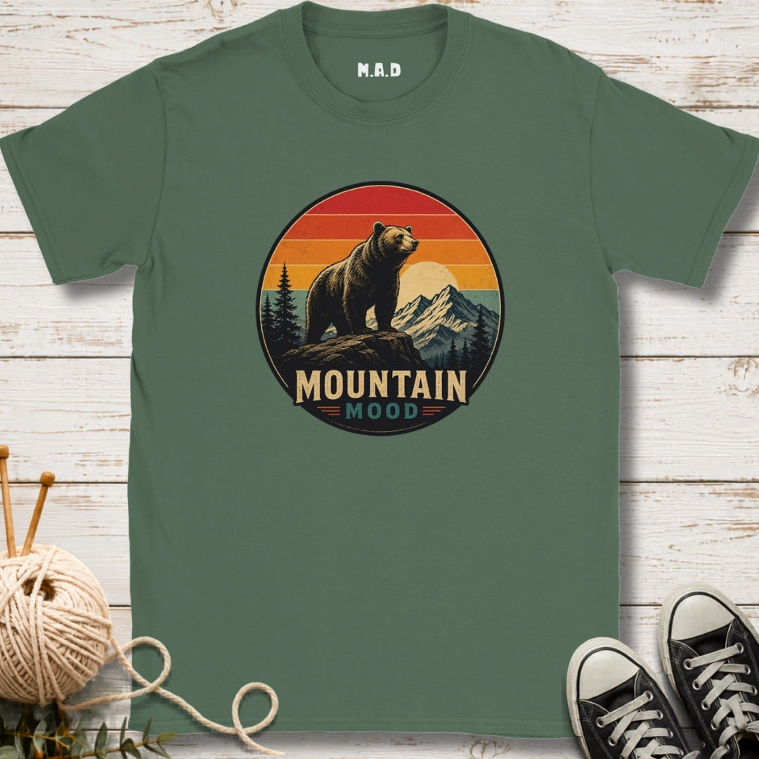 A sage green short-sleeve t-shirt with a circular retro graphic of a bear in front of mountains and a sunset, with the text 'MOUNTAIN MOOD'. The t-shirt is displayed flat on a white wood background with knitting needles and yarn on the left, and black sneakers on the right.