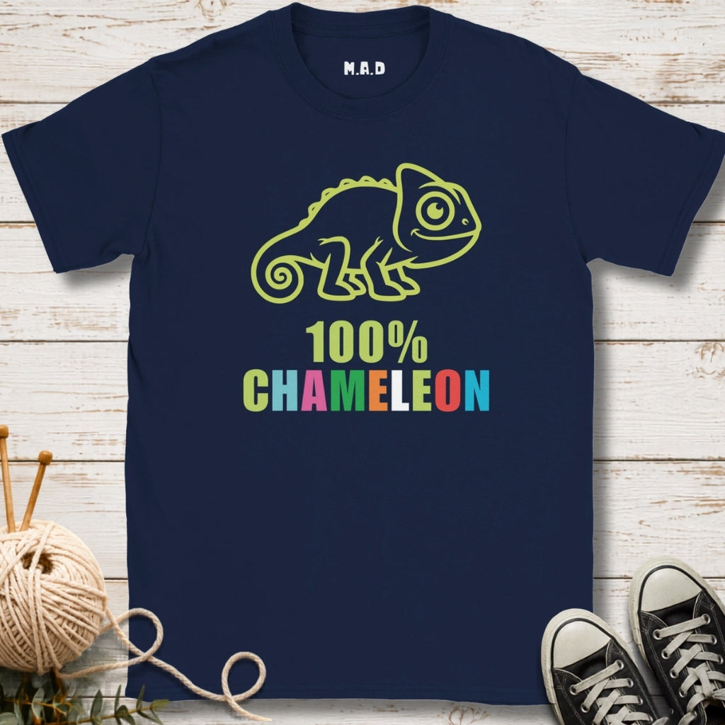 A navy blue short-sleeve t-shirt with a '100% CHAMELEON' graphic featuring a lime green chameleon and multicolored text, displayed on a white wood background with knitting needles, yarn, and black sneakers as props.
