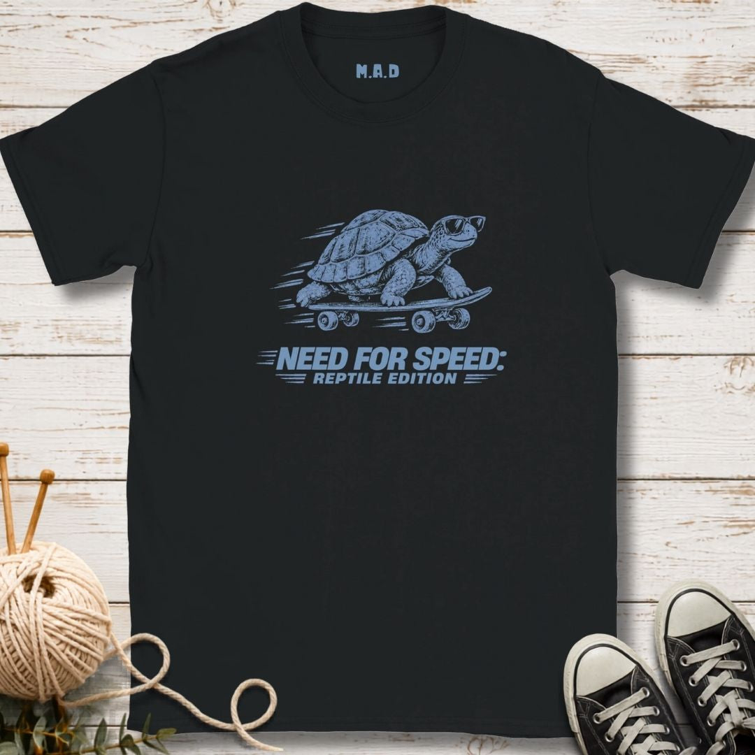A black short-sleeve graphic T-shirt laid flat on a whitewashed wood surface, with a blue turtle on a skateboard and the text 'NEED FOR SPEED: REPTILE EDITION' printed on the front. A ball of yarn, greenery, and black sneakers are placed around it as styling props.