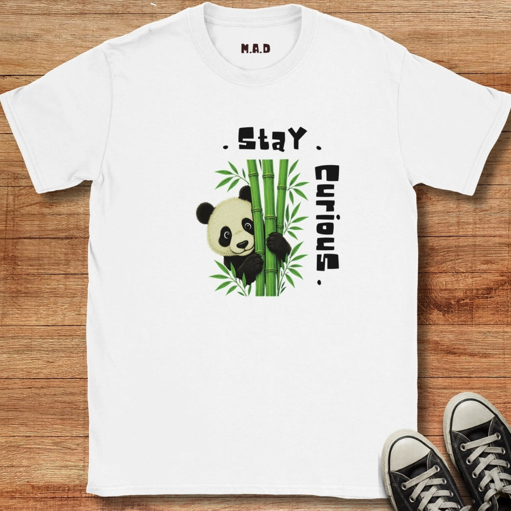 A white short-sleeve t-shirt with a graphic of a panda behind green bamboo and the text 'Stay Curious', laid flat on a wooden surface with black sneakers partially visible in the corner.