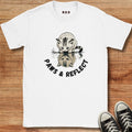 A white short-sleeve graphic T-shirt laid flat on a wooden surface, featuring a cat illustration above the words 'PAWS & REFLECT.' Black sneakers are partially visible at the bottom right as styling props.