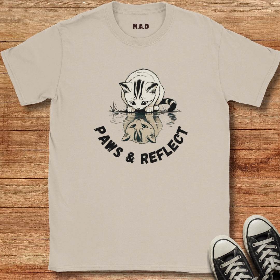A beige graphic t-shirt laid flat on a wooden background, featuring a black and white illustration of a cat looking at its reflection with the text 'PAWS & REFLECT'. Black sneakers are visible in the bottom right corner.