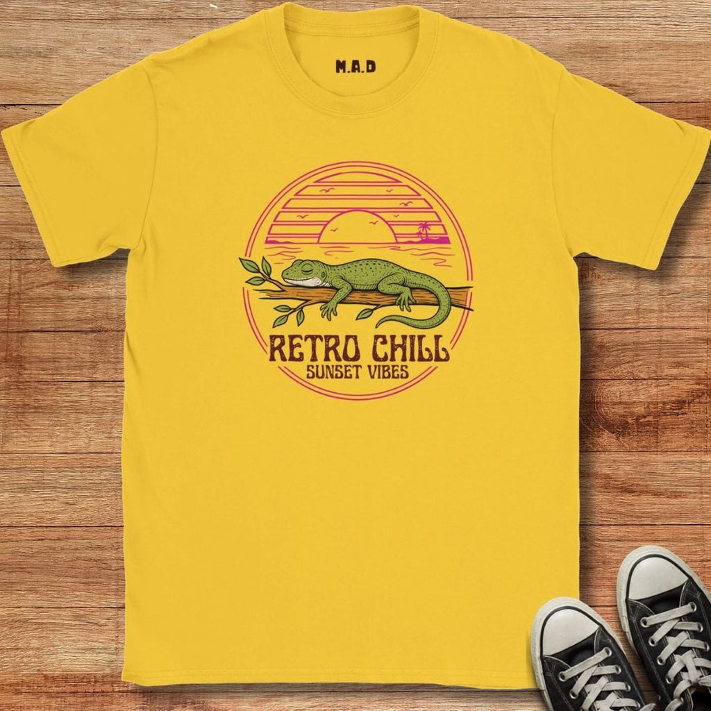 A bright yellow t-shirt with a retro-style lizard graphic and the text 'RETRO CHILL SUNSET VIBES' laid flat on a wooden background, with black sneakers partially visible in the bottom right corner.