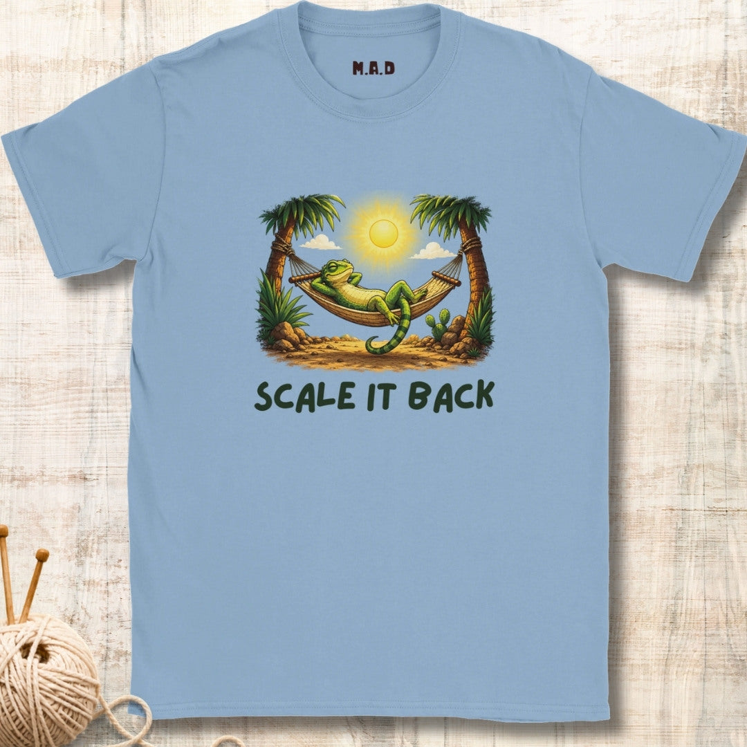A light blue crewneck t-shirt featuring a graphic of a green iguana relaxing in a hammock between two palm trees under a bright sun, with the text 'SCALE IT BACK' printed below. The shirt is displayed flat on a light wood background with a knitting needle and yarn prop in the lower-left corner.