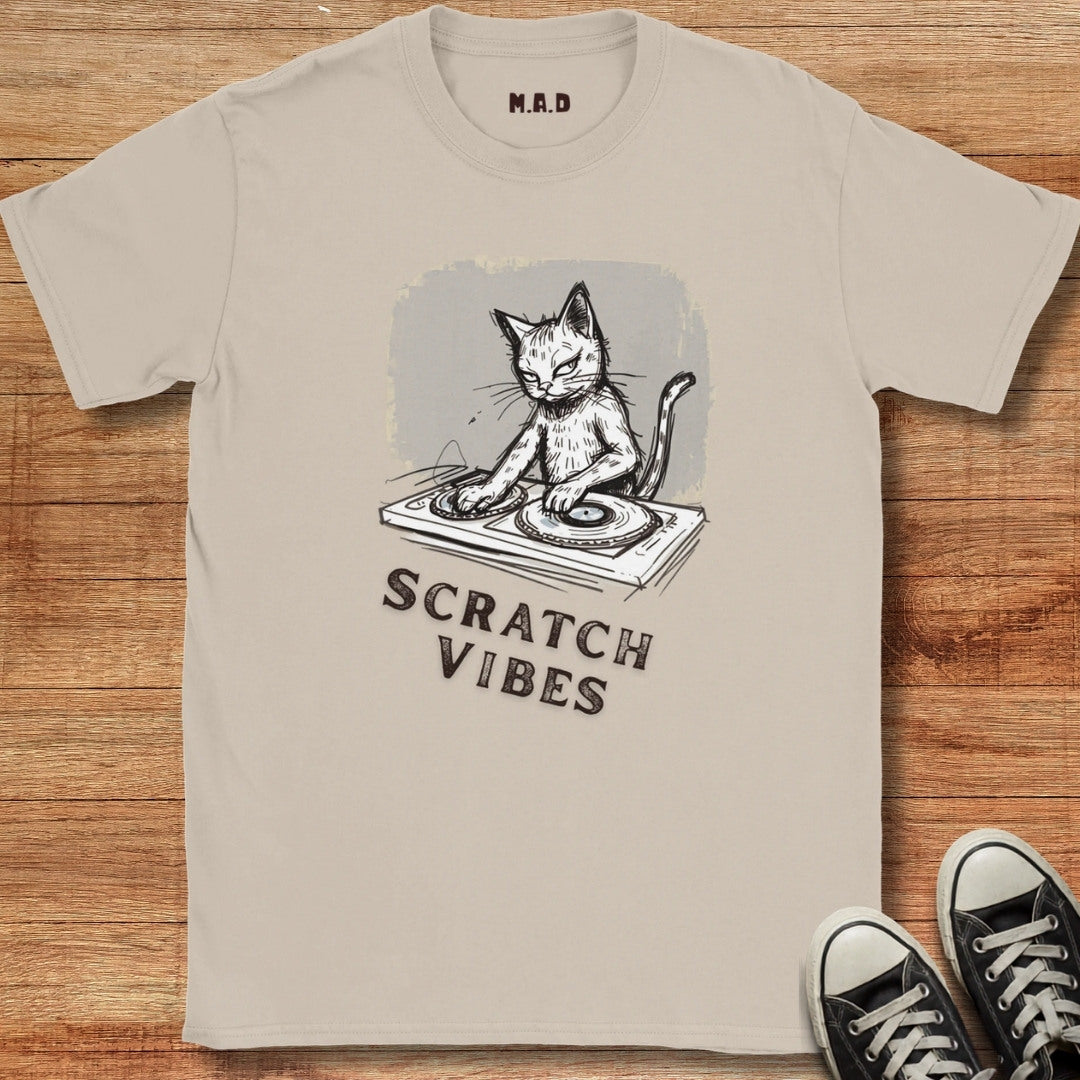 A beige graphic t-shirt laid flat on a wooden surface, featuring a black and white illustration of a cat DJing on a turntable with the text 'SCRATCH VIBES'. Black sneakers are visible in the bottom right corner.