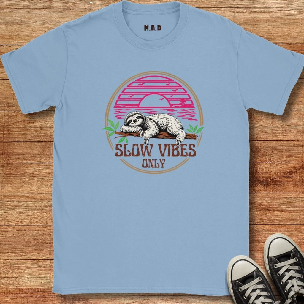 A light blue crewneck t-shirt with a retro-style sloth graphic on the front, laid flat on a wooden surface next to black sneakers.
