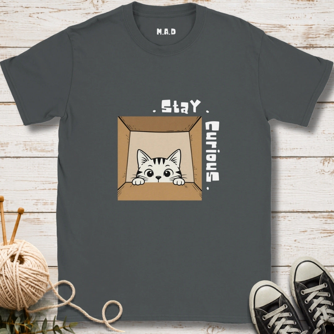 A dark gray t-shirt with a graphic of a curious cat peeking out of a cardboard box, accompanied by the text 'Stay Curious'. The shirt is displayed flat on a white wood background with knitting props and black sneakers.