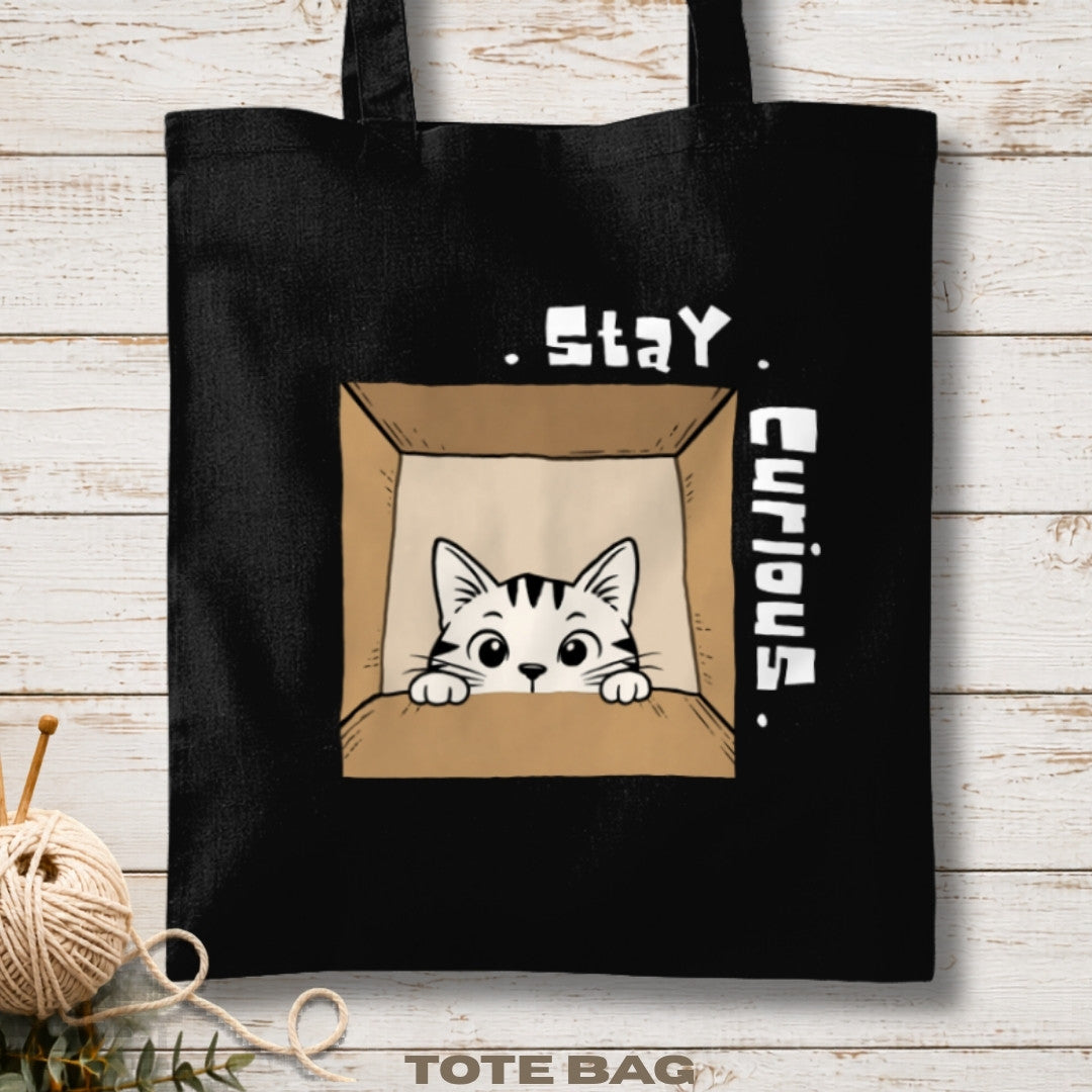 Black tote bag with a cat illustration and 'Stay Curious' text on a wooden surface.