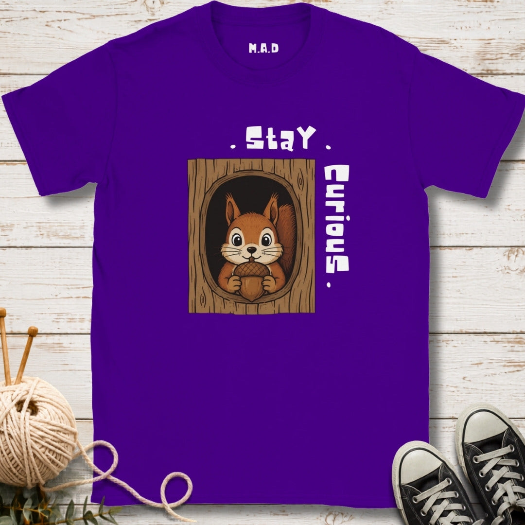 A purple short-sleeve t-shirt with a cartoon squirrel in a tree hole graphic and the text 'Stay Curious.' printed on the front, displayed on a whitewashed wood background with knitting yarn, shoes, and greenery as props.