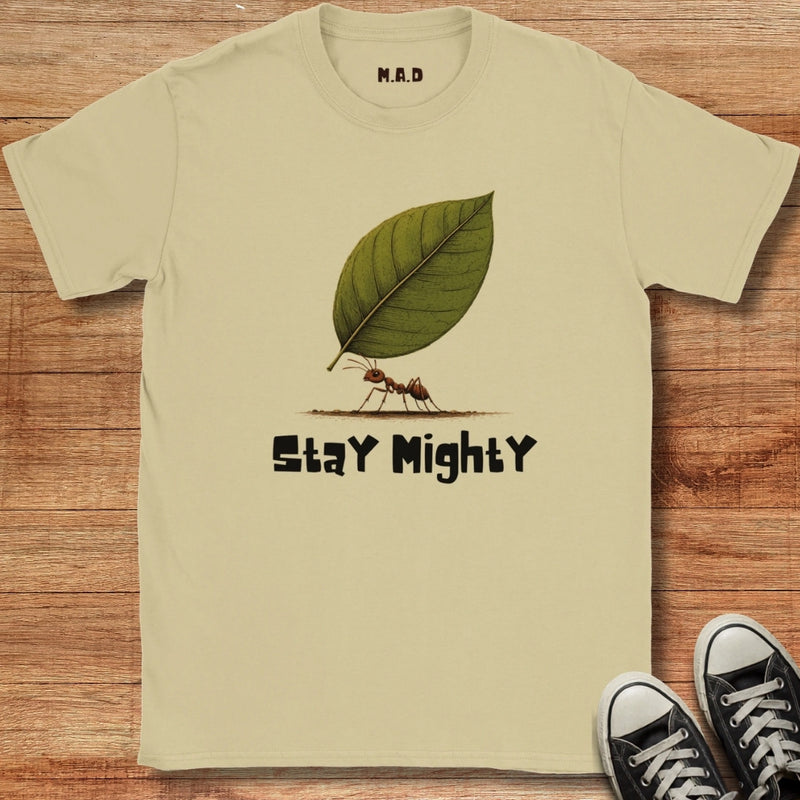 A beige graphic t-shirt laid flat on a wooden surface, featuring a green leaf and ant illustration with the text 'Stay Mighty'. Black sneakers are partially visible in the bottom right corner.