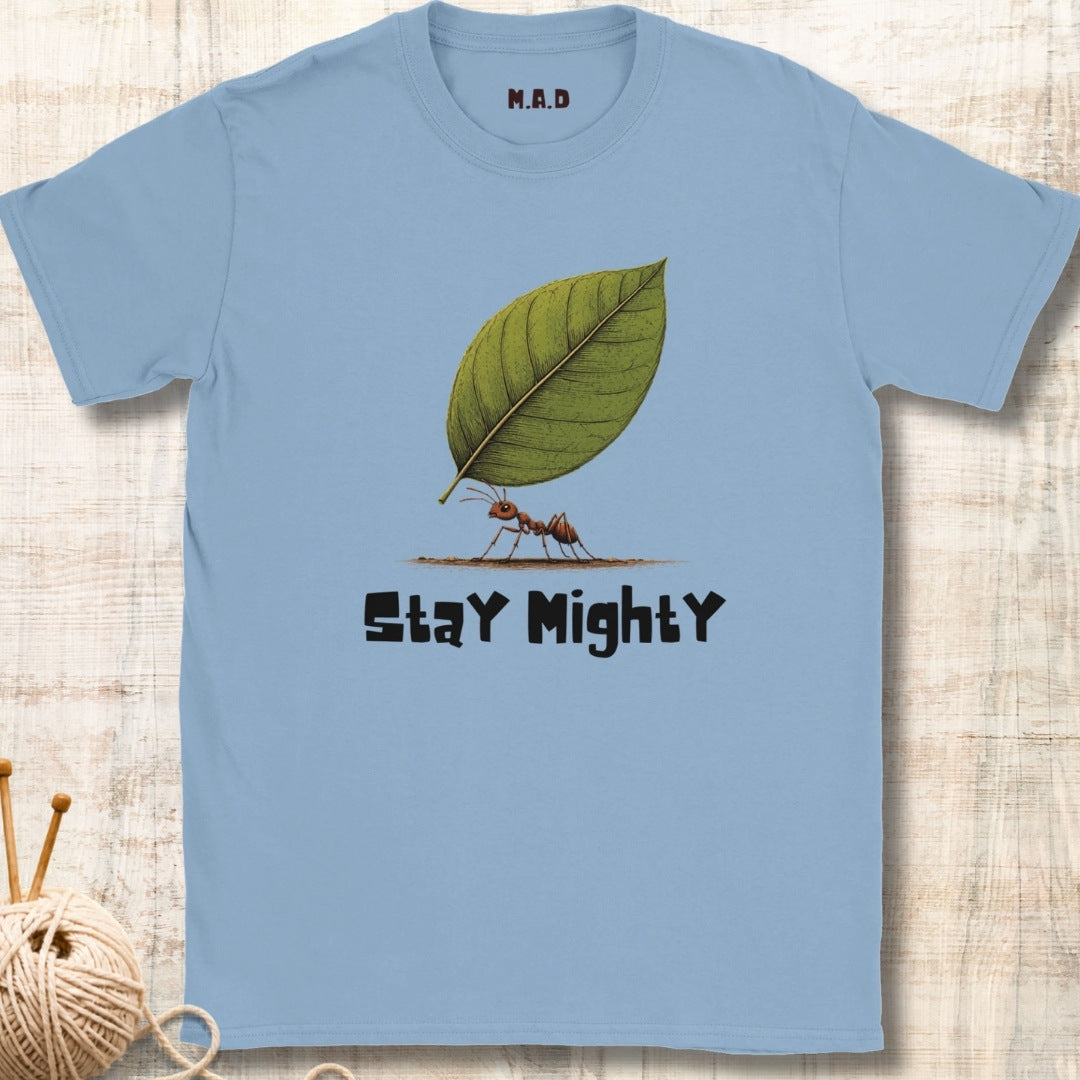 A light blue crewneck t-shirt featuring a graphic of an ant carrying a green leaf with the text 'Stay Mighty' printed below it. The shirt is displayed flat on a light wood background with a ball of yarn and knitting needles partially visible in the bottom-left corner.