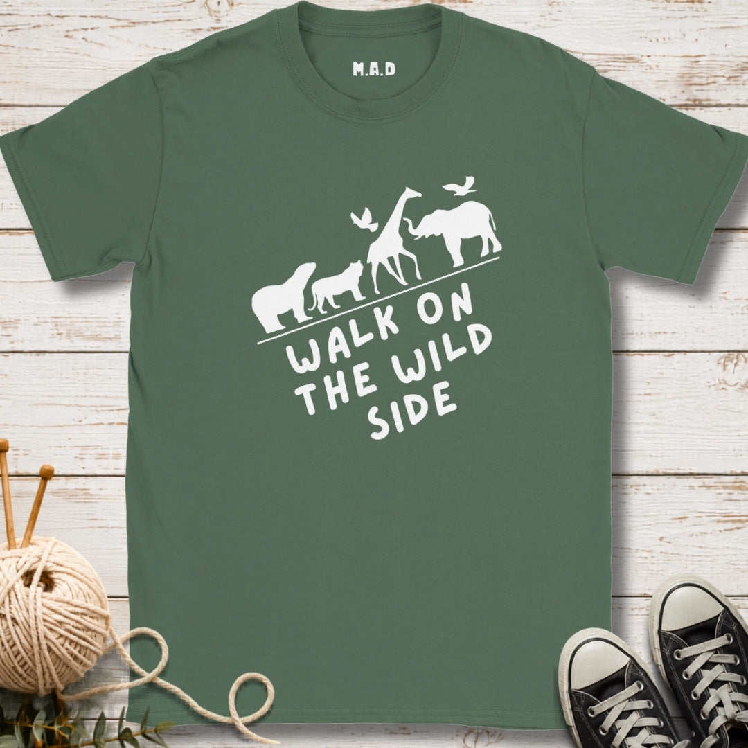 A sage green t-shirt with a white graphic of safari animals and the text 'WALK ON THE WILD SIDE' laid flat on a white wood background with knitting yarn and black sneakers as props.