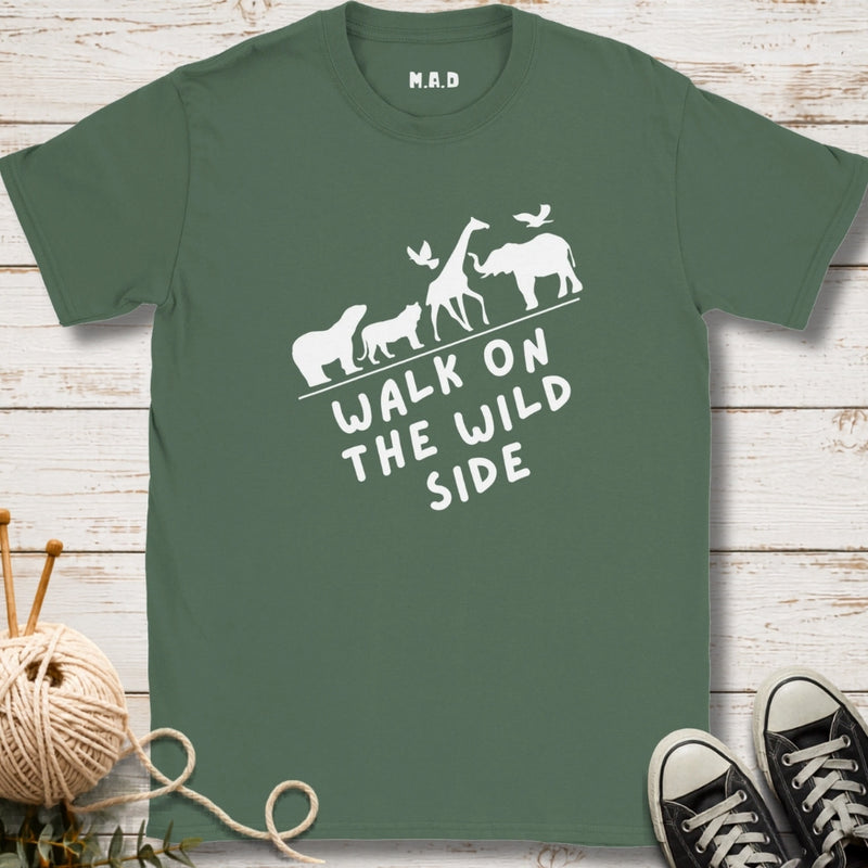 A sage green t-shirt with a white graphic of safari animals and the text 'WALK ON THE WILD SIDE' laid flat on a white wood background with knitting yarn and black sneakers as props.